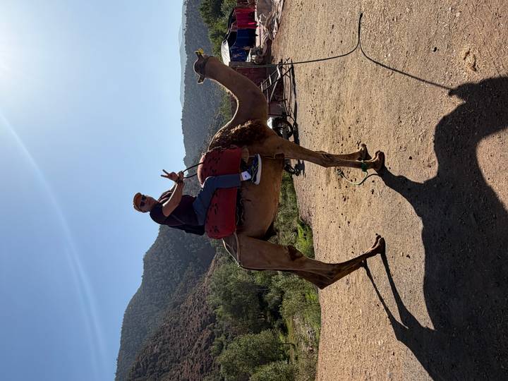 Man riding a camel, holding up peace sign, with mountain backdrop.