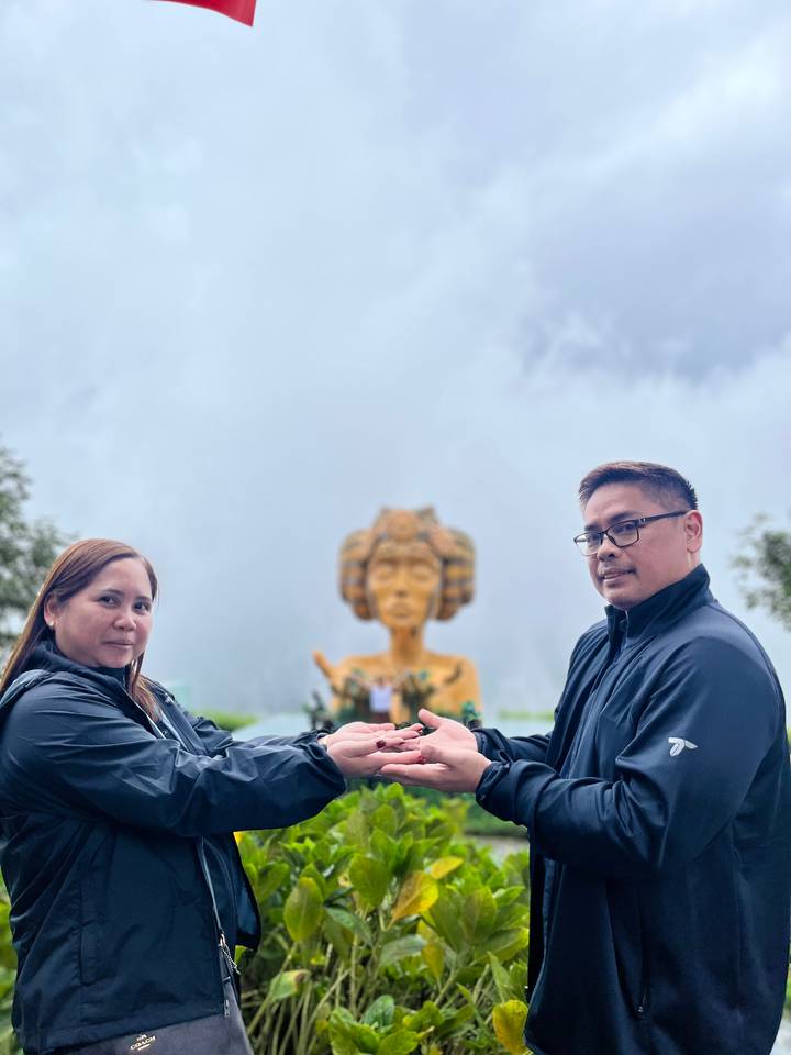 Couple holding hands in front of large golden statue of a female figure.