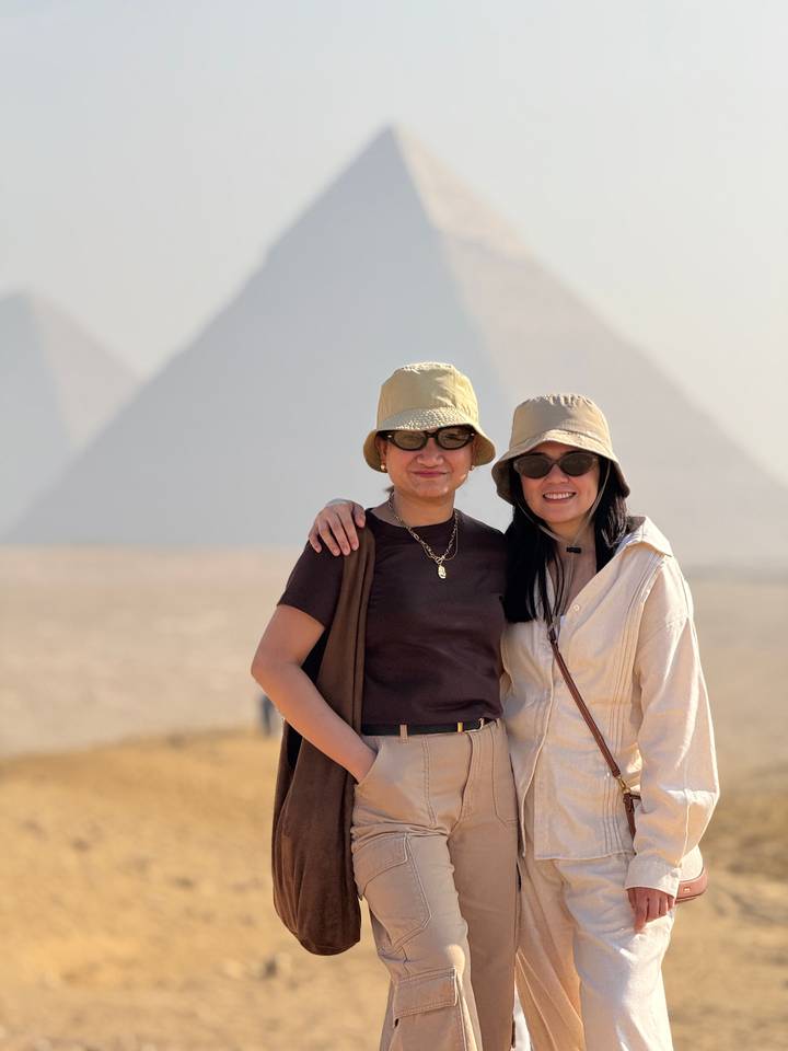 Twee mensen die poseren met piramides op de achtergrond, met hoeden en zonnebrillen op.