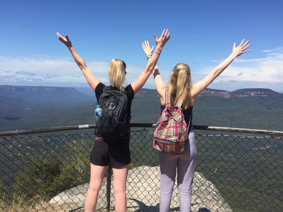 Two hikers raise their arms triumphantly at a mountain lookout over vast green valleys.