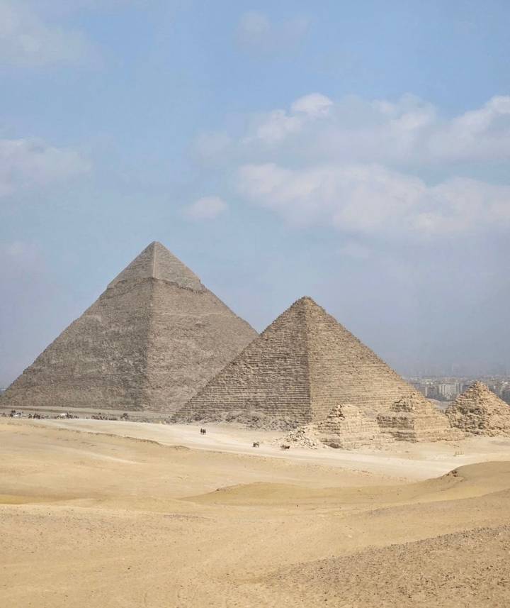 Two pyramids stand in the desert with a city view in the background.