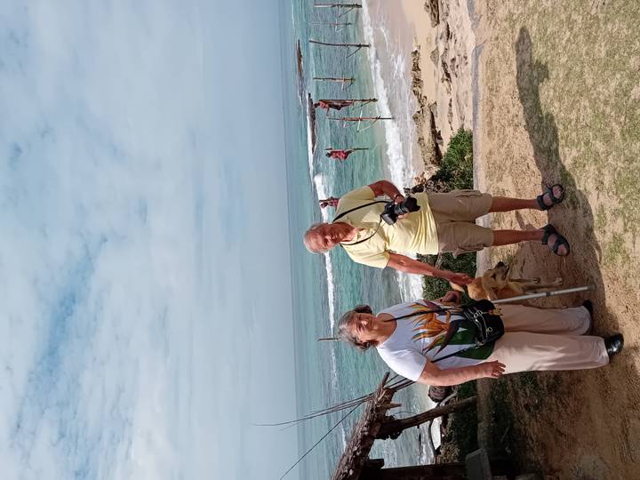 Oudere echtpaar en een hond op een strand met traditionele vissers op de achtergrond.