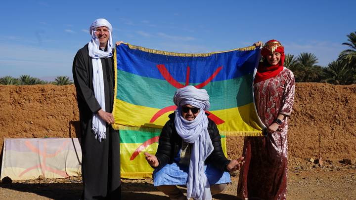 Tres personas con vestimenta tradicional sosteniendo una bandera colorida.