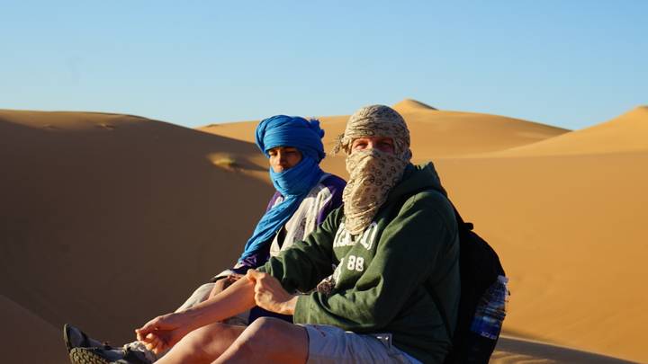 Two personas sentadas en dunas de arena del desierto usando pañuelos en la cabeza.