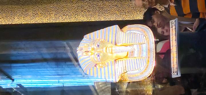 Masque doré de Toutânkhamon exposé dans un musée avec les reflets des visiteurs.