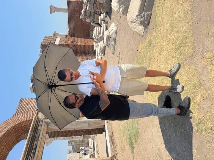 Zwei Männer betrachten ein Telefon unter einem Regenschirm an einer archäologischen Ausgrabungsstätte.