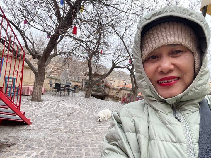 Person taking selfie in a cobblestone street with a dog lying nearby.