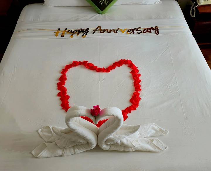 Bed decorated with a heart made of rose petals and towel swans for an anniversary celebration.