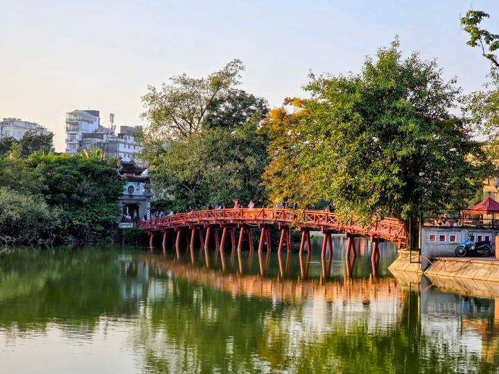 Le pont rouge emblématique de Hanoï au-dessus d'un lac calme avec des arbres verts et des bâtiments.