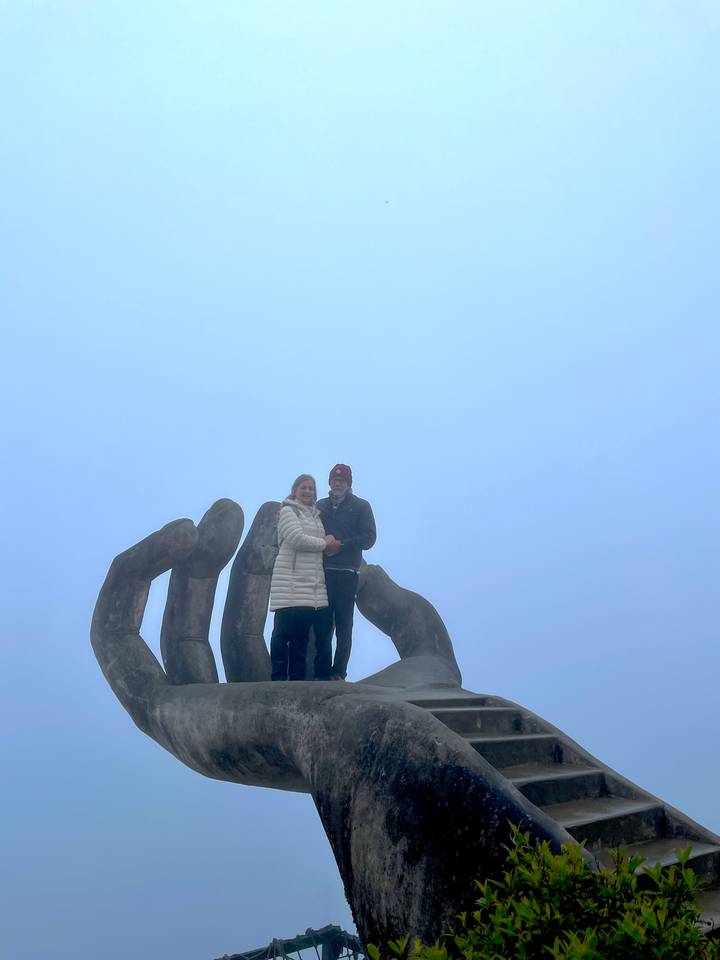 Dos personas paradas sobre una gran escultura de mano en un ambiente brumoso.