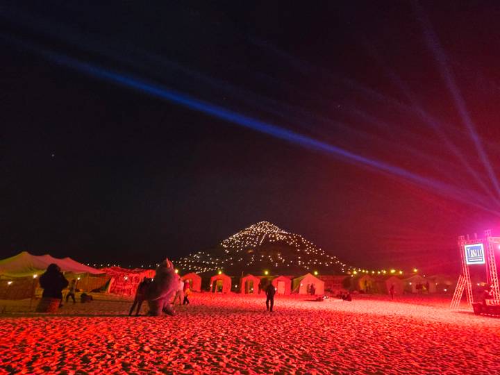 Night view with tents and people illuminated by red lights, with a lit mountain in the background.