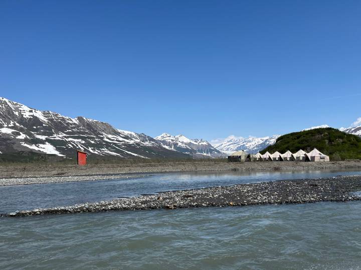 Tiendas de campaña con montañas y un río visible al fondo bajo un cielo azul despejado.