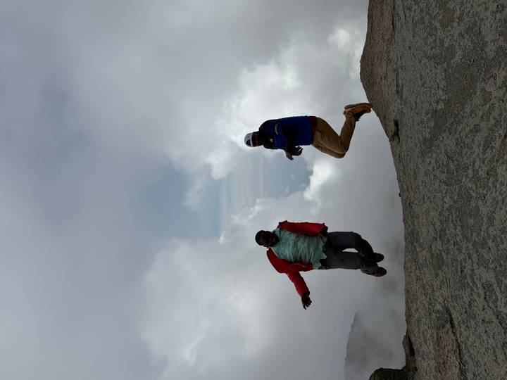 Dos personas saltando con alegría en la cima rocosa de una montaña con nubes.