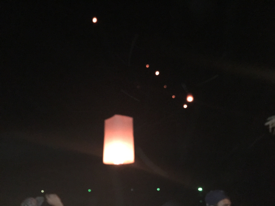 Blurred night photo of a glowing sky lantern rising with several others in the dark sky.