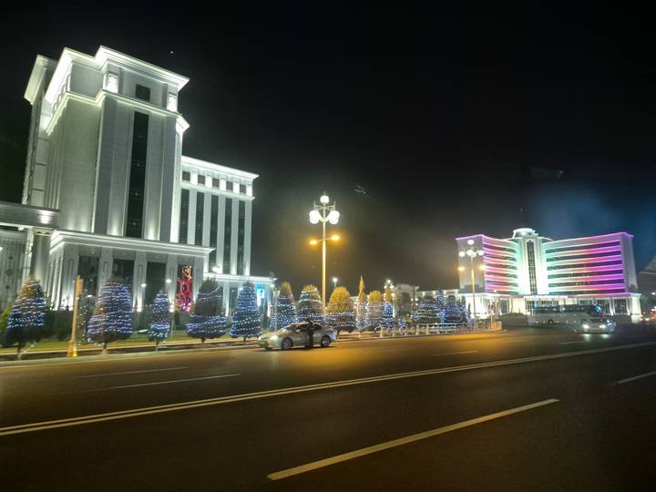 Nighttime cityscape with brightly lit modern buildings and a road with traffic.