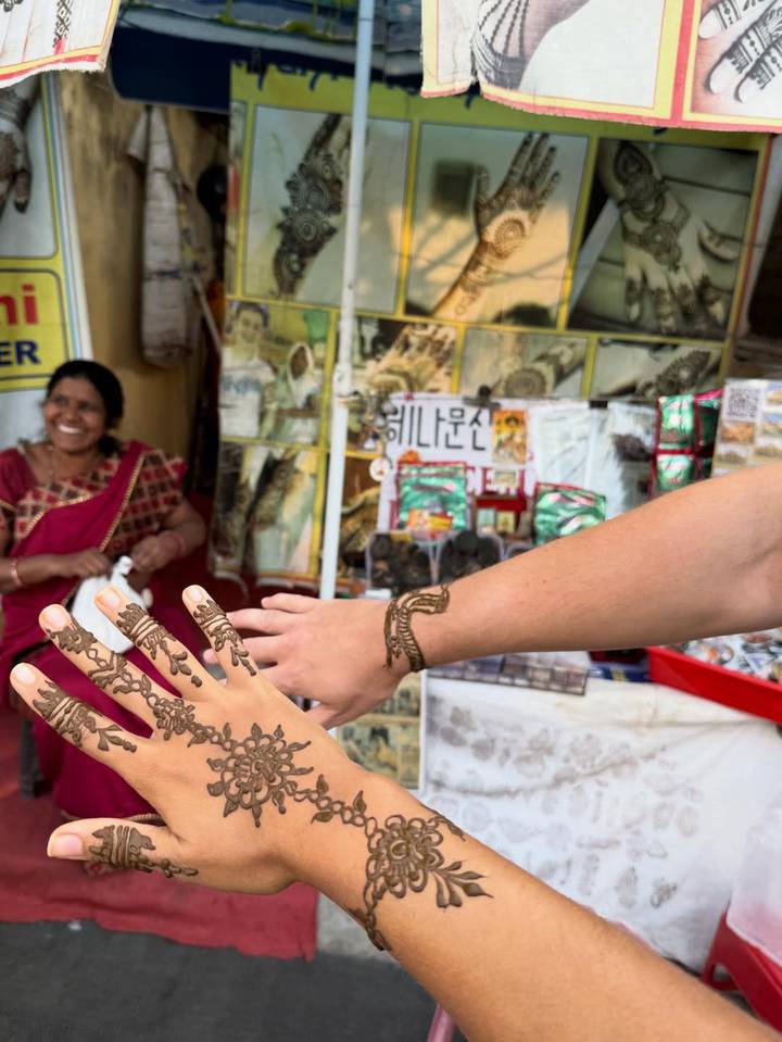 Una persona mostrando henna en su mano con un comerciante sonriente en el fondo.
