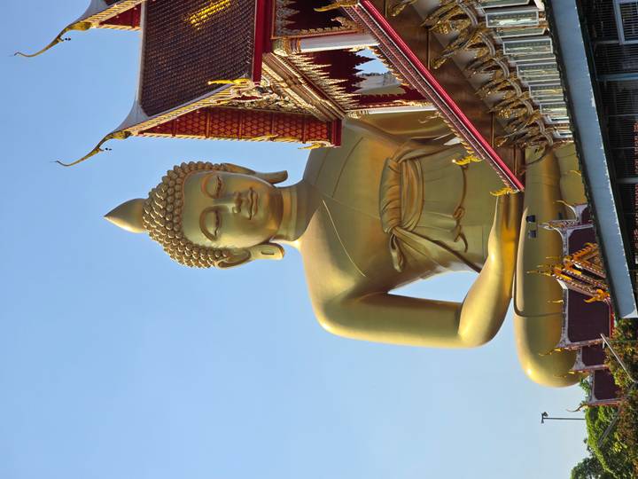Gran estatua dorada de Buda junto a un templo bajo un cielo azul.
