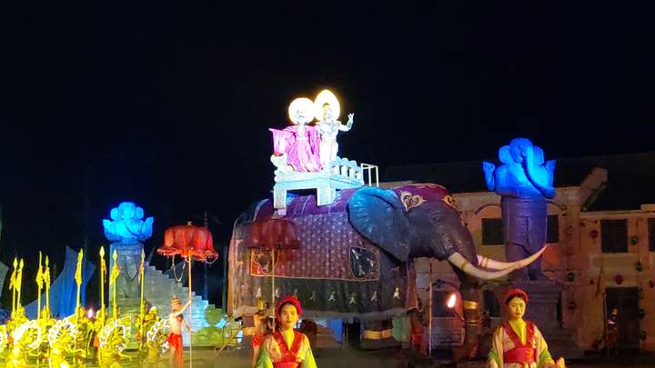 Actuación con personas en vestimenta tradicional y una carroza temática de elefante por la noche.