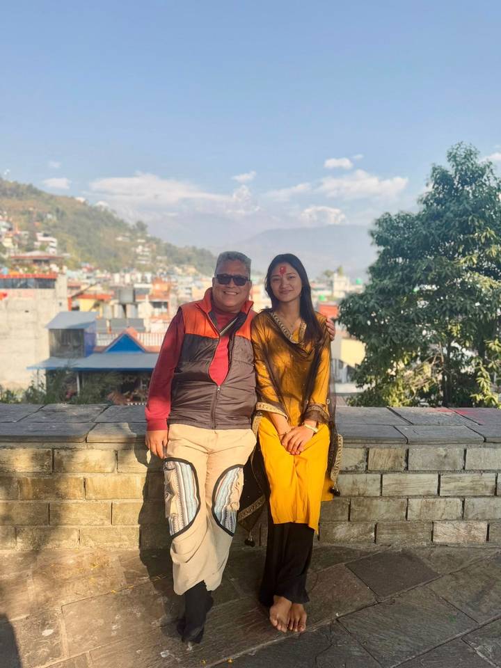 Dos personas sentadas en un banco, con un paisaje urbano de Nepal de fondo.
