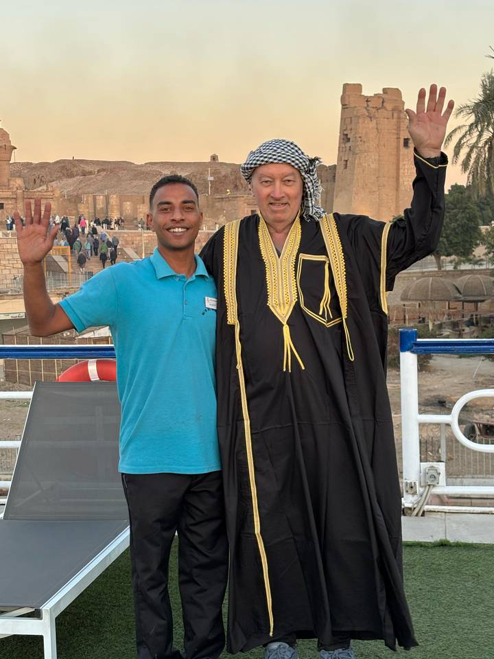Dos hombres posando con vestimenta tradicional en un barco con ruinas antiguas al fondo.