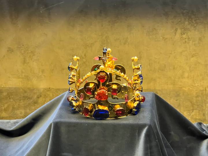 Corona dorada con joyas exhibida sobre un pedestal cubierto con tela.