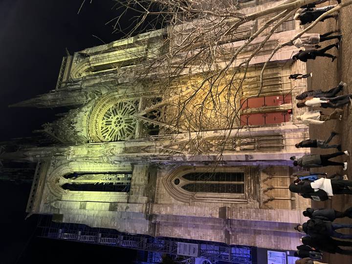 Fachada de iglesia gótica con personas caminando enfrente, iluminada de noche.
