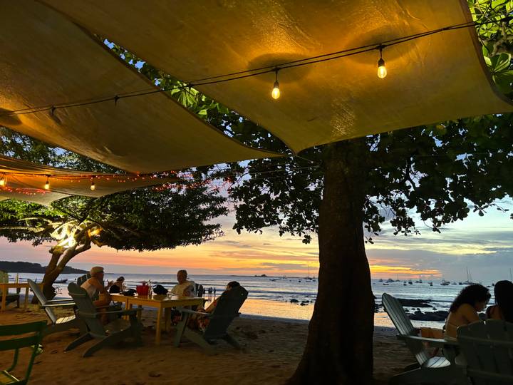 Des personnes dînant en plein air sous des guirlandes lumineuses avec vue sur le coucher de soleil.