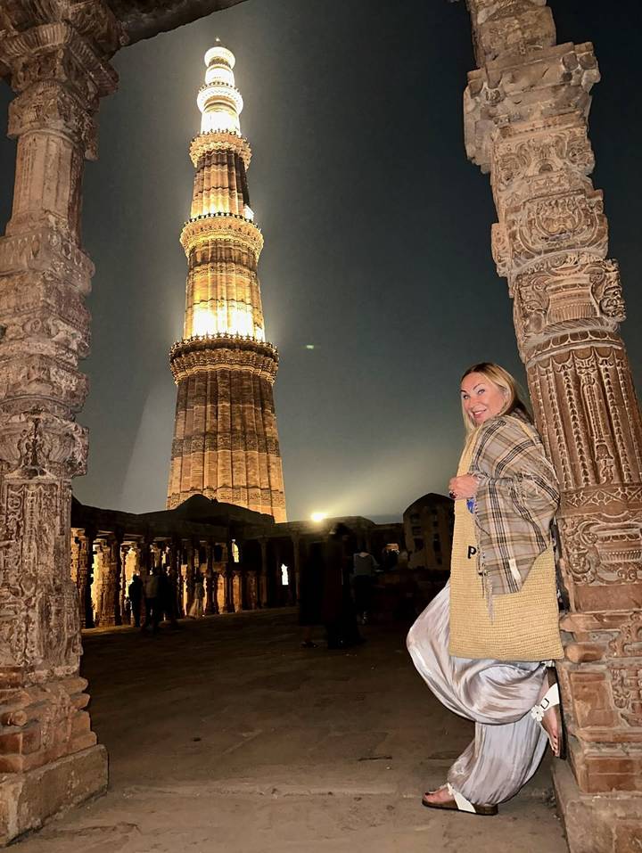 Persona de pie en un monumento antiguo durante la noche, con un pilar iluminado en el fondo.