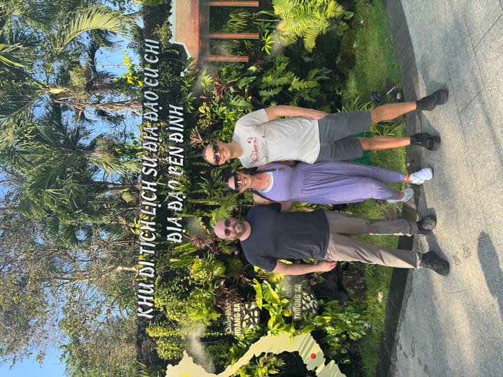 Trois personnes posant devant un panneau avec des plantes tropicales derrière elles.