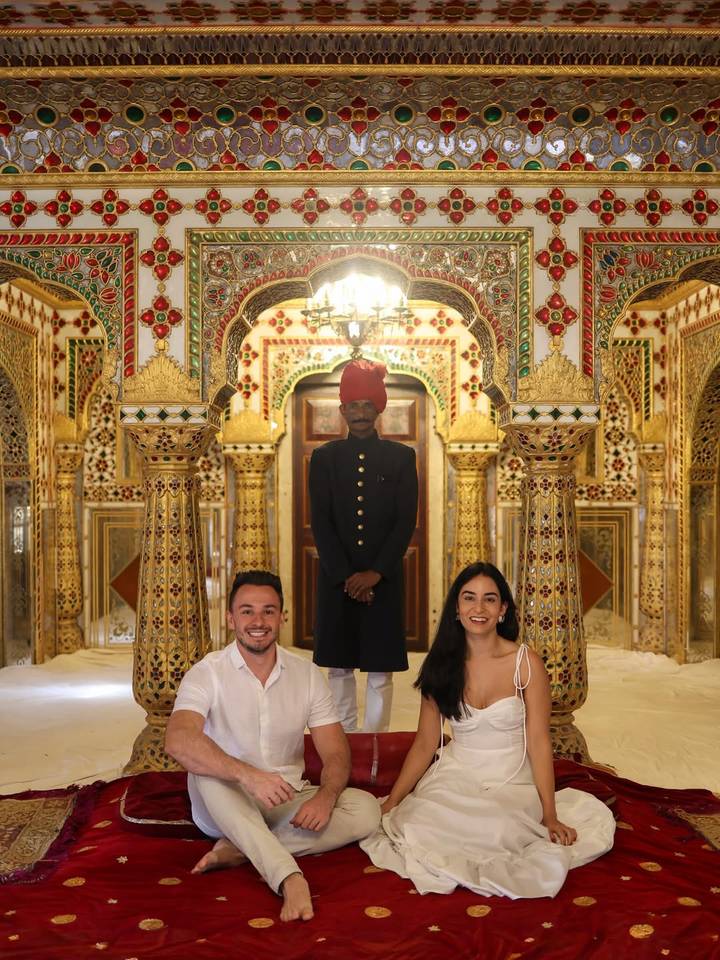 Tres personas en un interior ornamentalmente decorado con un guardia vestido con atuendo cultural.