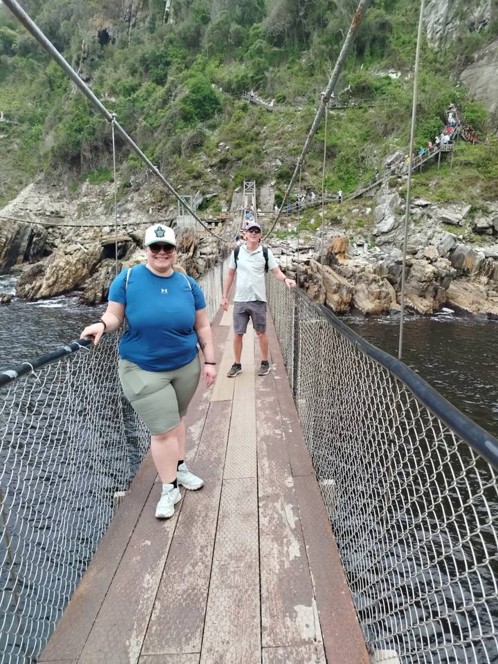 Zwei Personen überqueren eine Hängebrücke über einen Fluss in einer üppigen Gegend.