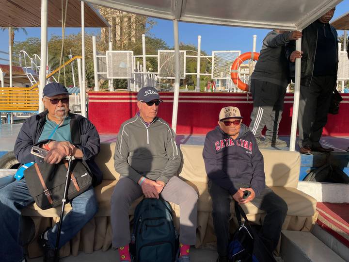 Three older men sitting on a boat deck under sunlight.