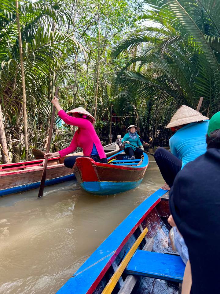 Group of people in traditional boats navigating a narrow waterway surrounded by trees.