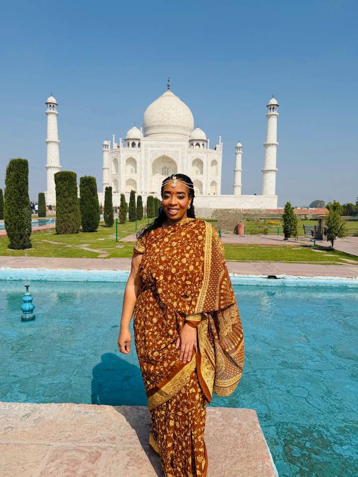 Full-body portrait of woman in ornate sari standing before turquoise pool and Taj Mahal.