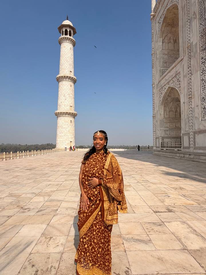 Woman in patterned sari standing on the marble terrace beside a Taj Mahal minaret under clear sky.