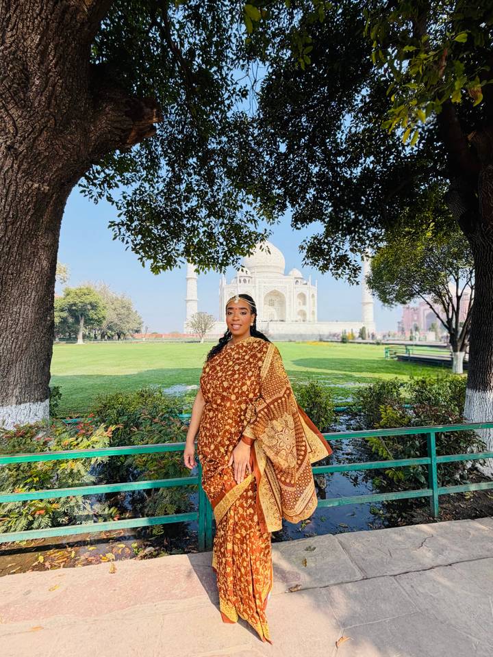 Woman in sari framed by leafy trees with distant view of the Taj Mahal across lush grounds.