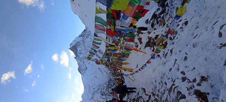 Prayer flags flutter against a snowy mountain backdrop with trekkers gathering nearby.