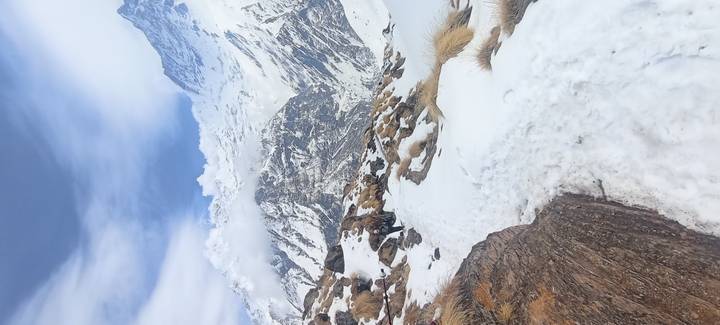 Snowy trail winding through tussock grasses toward towering Himalayan peaks under patchy clouds.