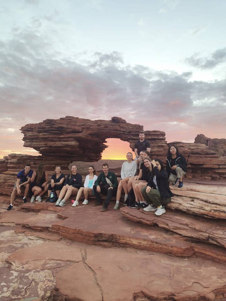 Group of travellers posing on a red rock ledge framed by a natural sandstone arch at sunset.