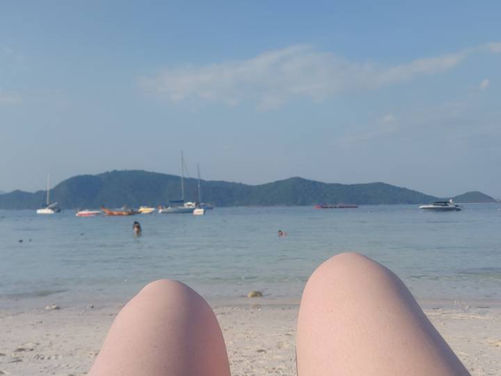 Beach view from the perspective of someone lying down with knees in foreground, boats and islands beyond.