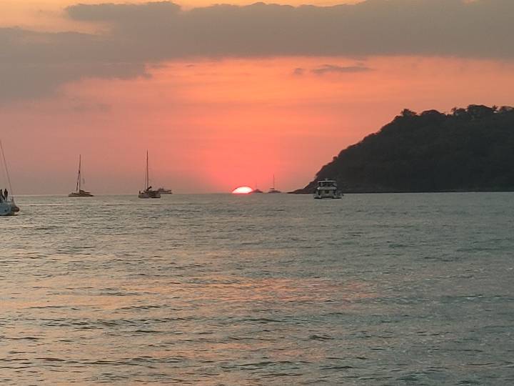 Orange sun setting behind a wooded headland with sailboats scattered on calm water.