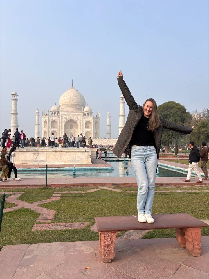 A joyful traveller raises her arms in front of the Taj Mahal with visitors milling around the reflective pools.