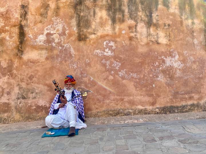 A traditionally dressed Rajasthani musician sits against a weathered wall playing a string instrument.