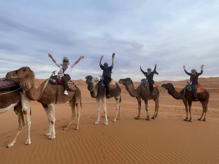 Five travellers ride camels across rolling Sahara dunes raising their arms in excitement.