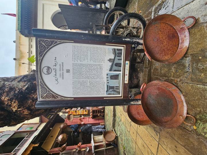 Informative signboard for Medersa Seffarine displayed on a rainy street with copper trays at the base.