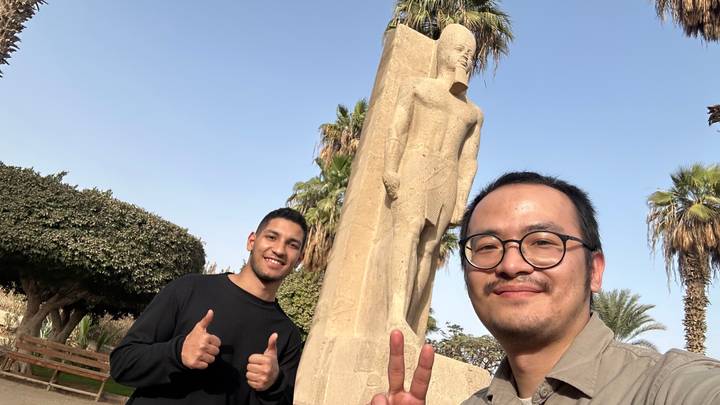 Two friends flash thumbs-up beside an ancient limestone statue amid palm trees.