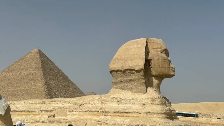 Side profile of the Great Sphinx with the Pyramid of Khafre rising behind it under a blue sky.
