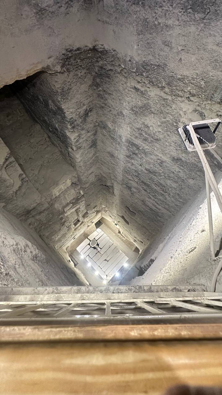 Dimly lit sloping interior passageway inside an ancient pyramid with rough stone walls and a ventilator fan.