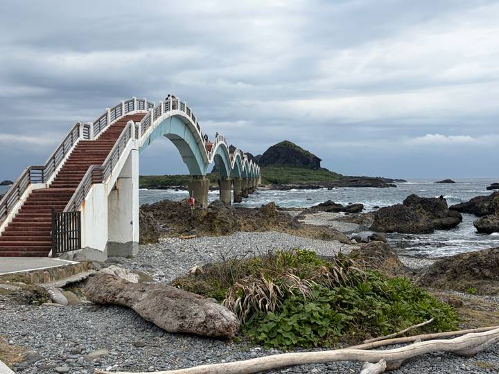 Arched Sanxiantai footbridge spans rocky shoreline toward a small offshore island under gray skies.