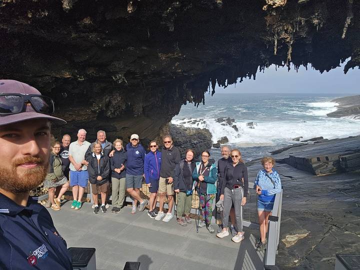 Large tour group and guide take a selfie under a rugged sea cave with waves crashing on rocks behind.